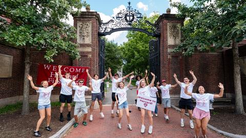 Student Orientation leaders jump up in the air as they welcome first-year students during move-in in the front of Johnston Gate.